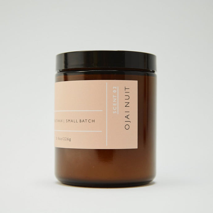 Side view of Roen's Ojai Nuit scented candle in an amber brown glass jar with a black lid and pink label.